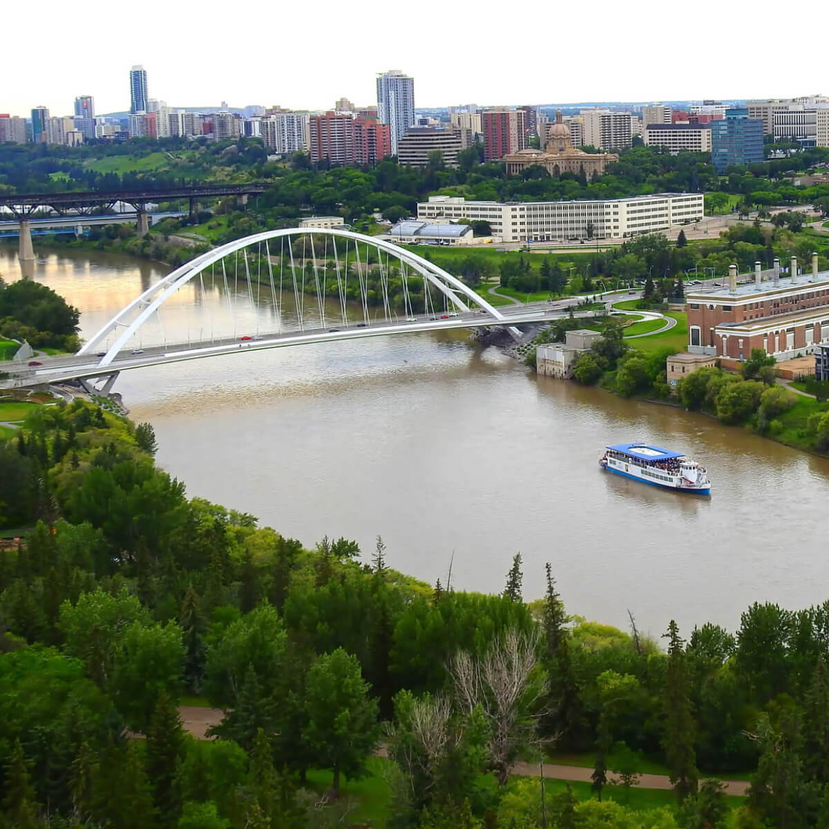 A landscape photo of Edmonton's river valley showing trees, the river, a ferry, a bridge, and buildings in the background